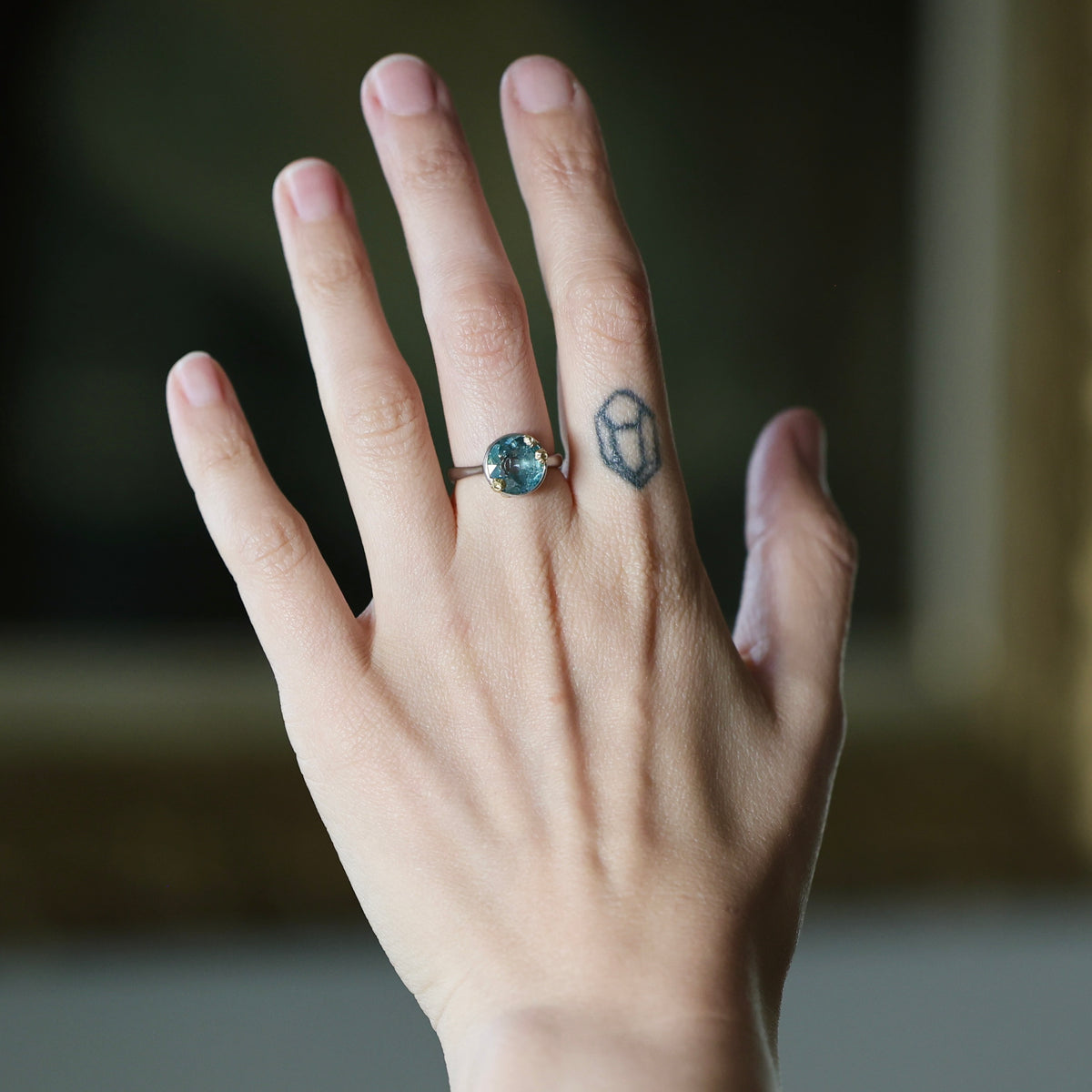 A close-up of a hand wearing a silver ring with a round blue gemstone, with a small geometric tattoo on the middle finger, set against a softly blurred dark background.