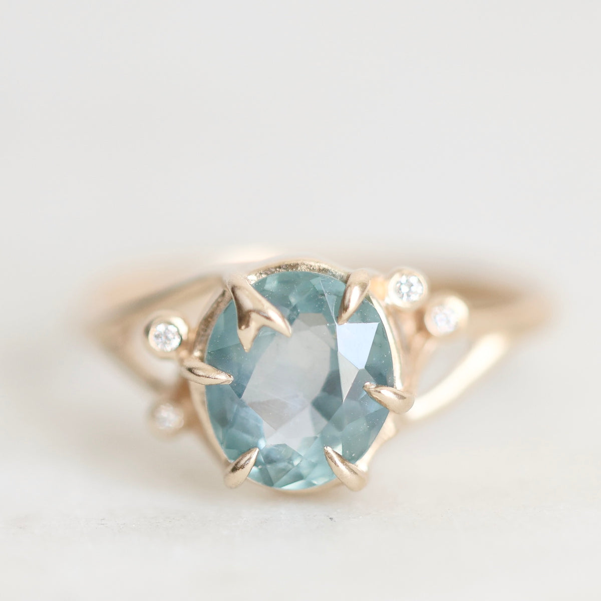 A gold ring featuring a large oval light blue gemstone in a prong setting, accented by three small round clear stones on the shoulders, shown against a soft light gray background.