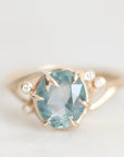 A gold ring featuring a large oval light blue gemstone in a prong setting, accented by three small round clear stones on the shoulders, shown against a soft light gray background.