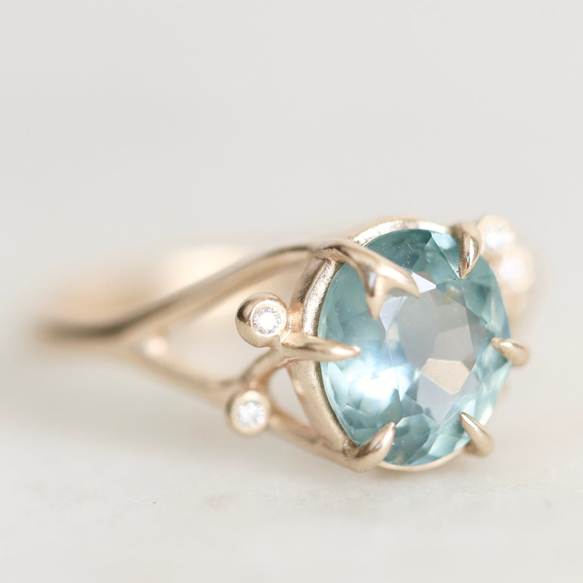 A close-up shot of a gold ring featuring a large oval-cut light blue gemstone, accented by two small round-cut diamonds on the band, set against a soft, light gray background.