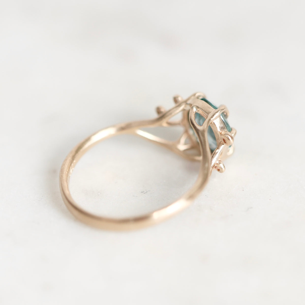A close-up shot of a vintage-style ring featuring a gold-toned band and a bezel-set teal gemstone, resting on a light marble surface.