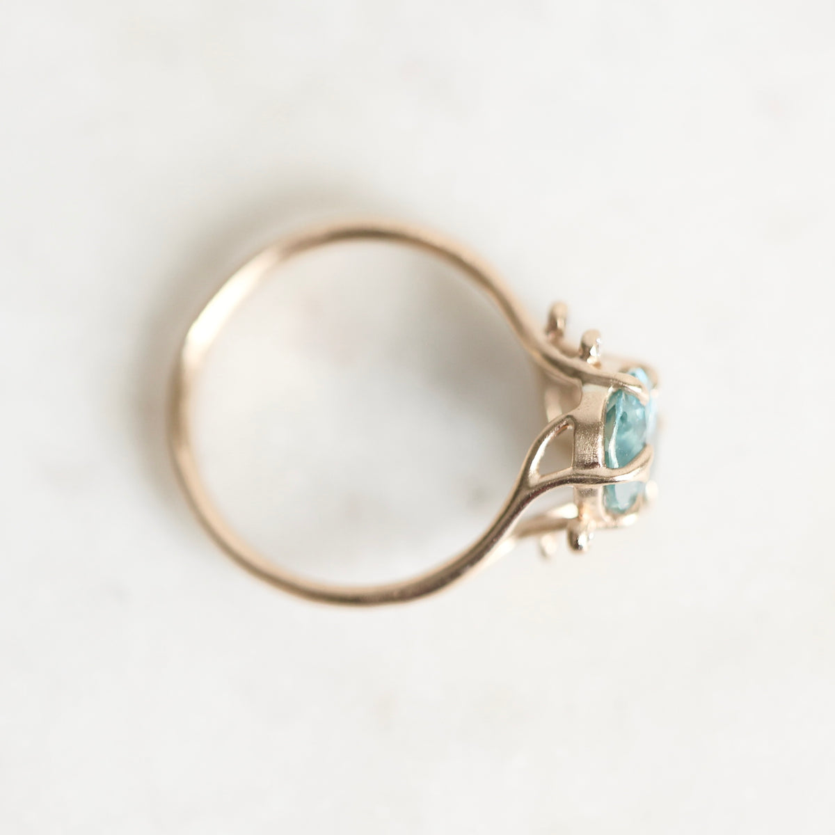 A gold ring with two light blue oval gemstones, photographed from an overhead angle on a light, off-white textured surface.