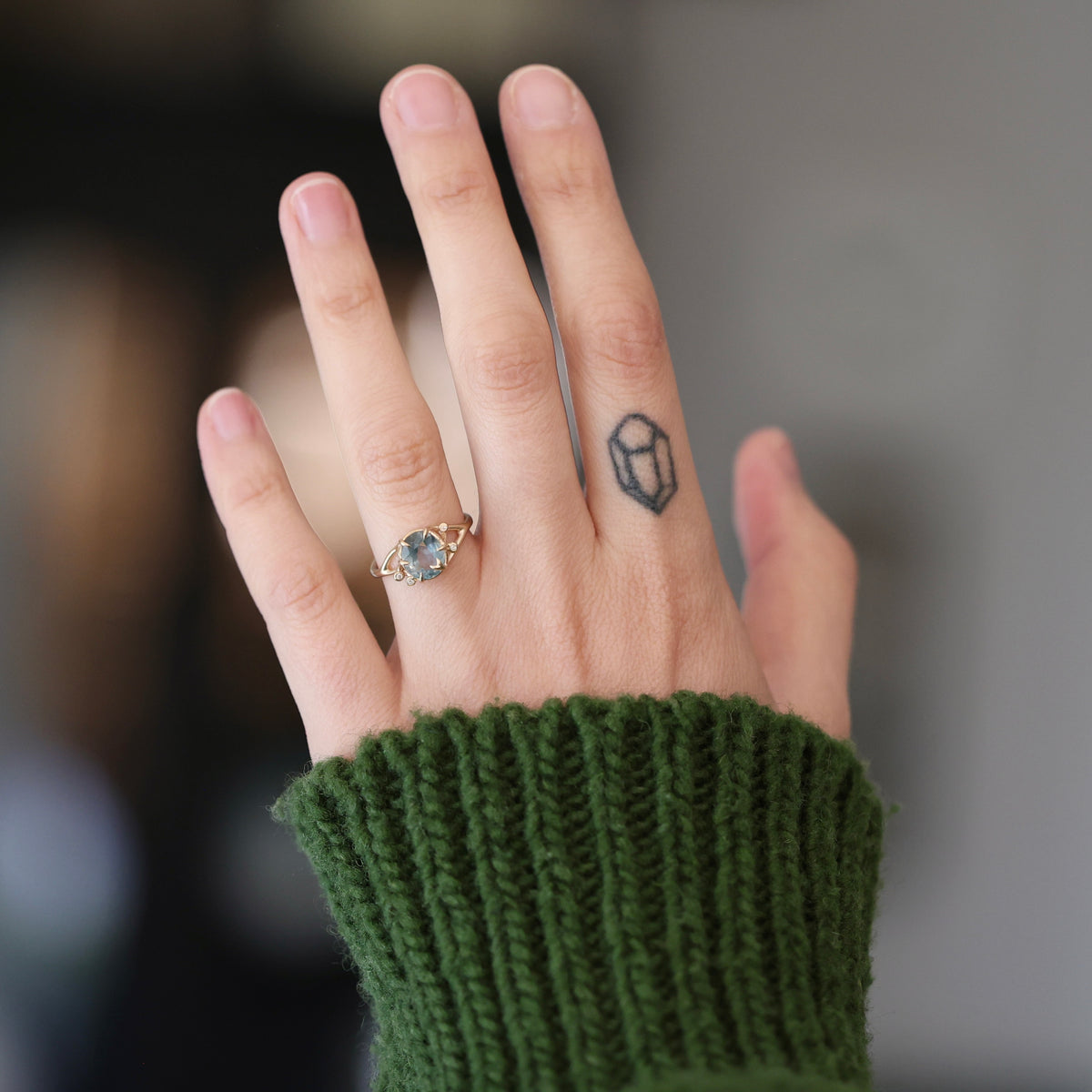 A close-up of a hand wearing a delicate gold ring with a small oval blue gemstone, styled with a dark green ribbed knit sweater sleeve. The background is softly blurred.
