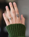 A close-up of a hand wearing a delicate gold ring with a small oval blue gemstone, styled with a dark green ribbed knit sweater sleeve. The background is softly blurred.