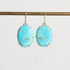 A pair of oval turquoise drop earrings with gold-toned prong settings and hooks, hanging from a gold rod against a light gray background.