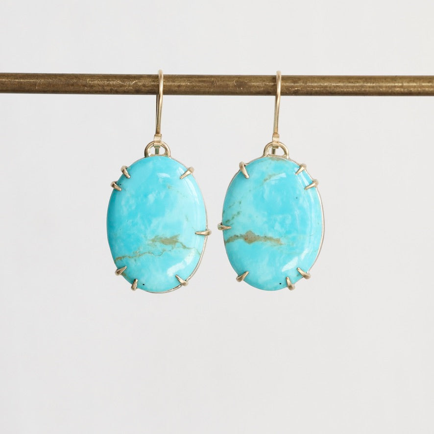 A pair of oval turquoise drop earrings with gold-toned prong settings and hooks, hanging from a gold rod against a light gray background.