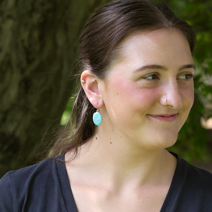 A woman outdoors wearing a single turquoise oval drop earring, with a blurred green tree background.