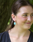 A woman outdoors wearing a single turquoise oval drop earring, with a blurred green tree background.