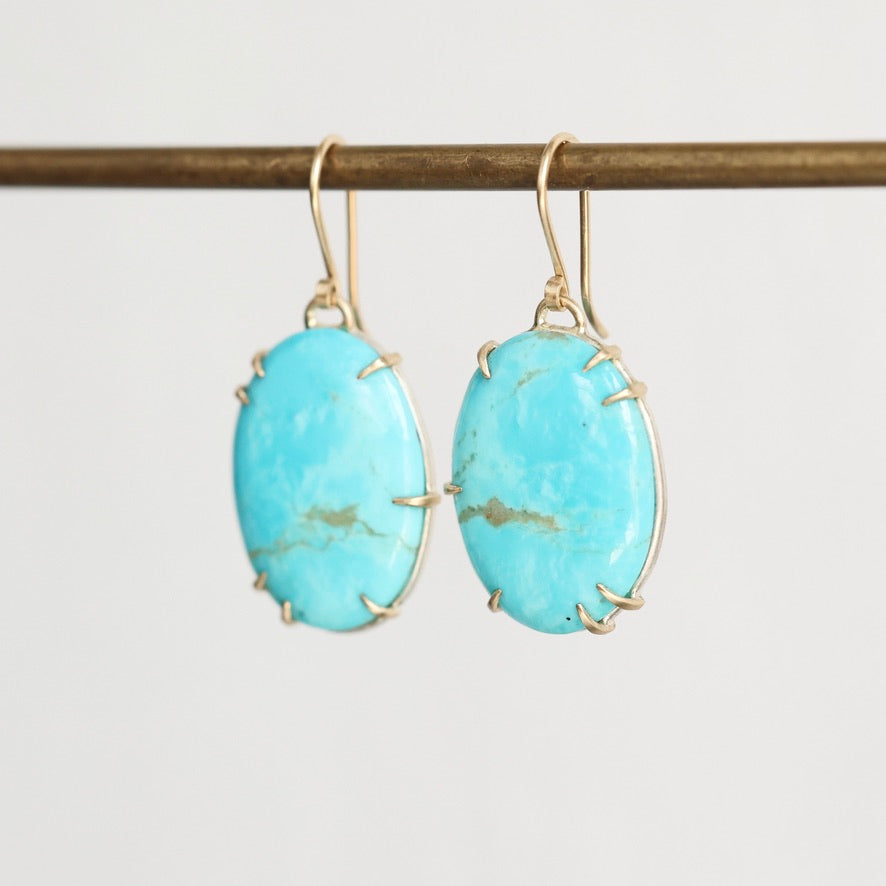 A pair of oval turquoise drop earrings with gold wire wrap settings and gold fishhook ear wires, hanging from a gold-colored horizontal bar against a light gray background.