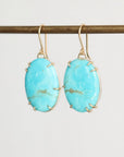A pair of oval turquoise drop earrings with gold wire wrap settings and gold fishhook ear wires, hanging from a gold-colored horizontal bar against a light gray background.