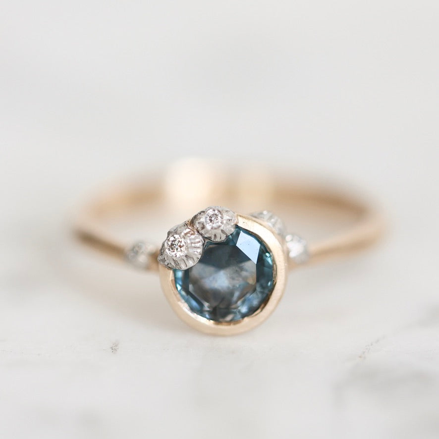 A gold ring featuring a round dark blue gemstone in a bezel setting, accented with small clear stones along the band, photographed on a clean white background.