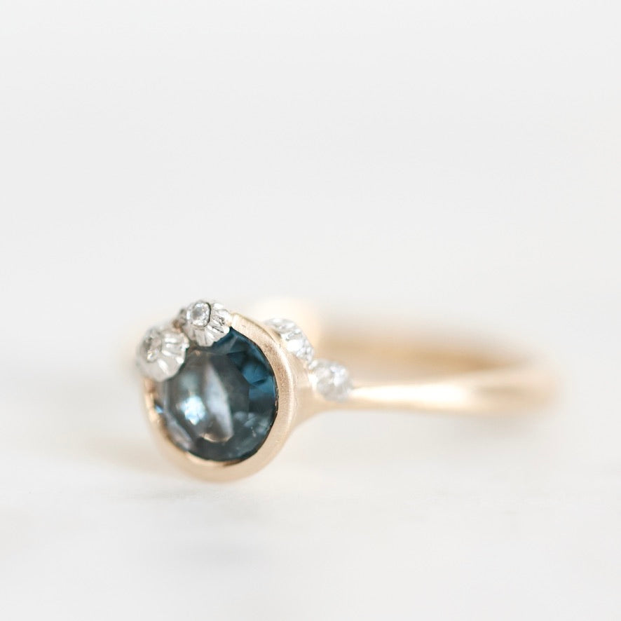 A gold ring featuring a round dark blue gemstone in a bezel setting, accented with small clear stones along the band, photographed on a clean white background.