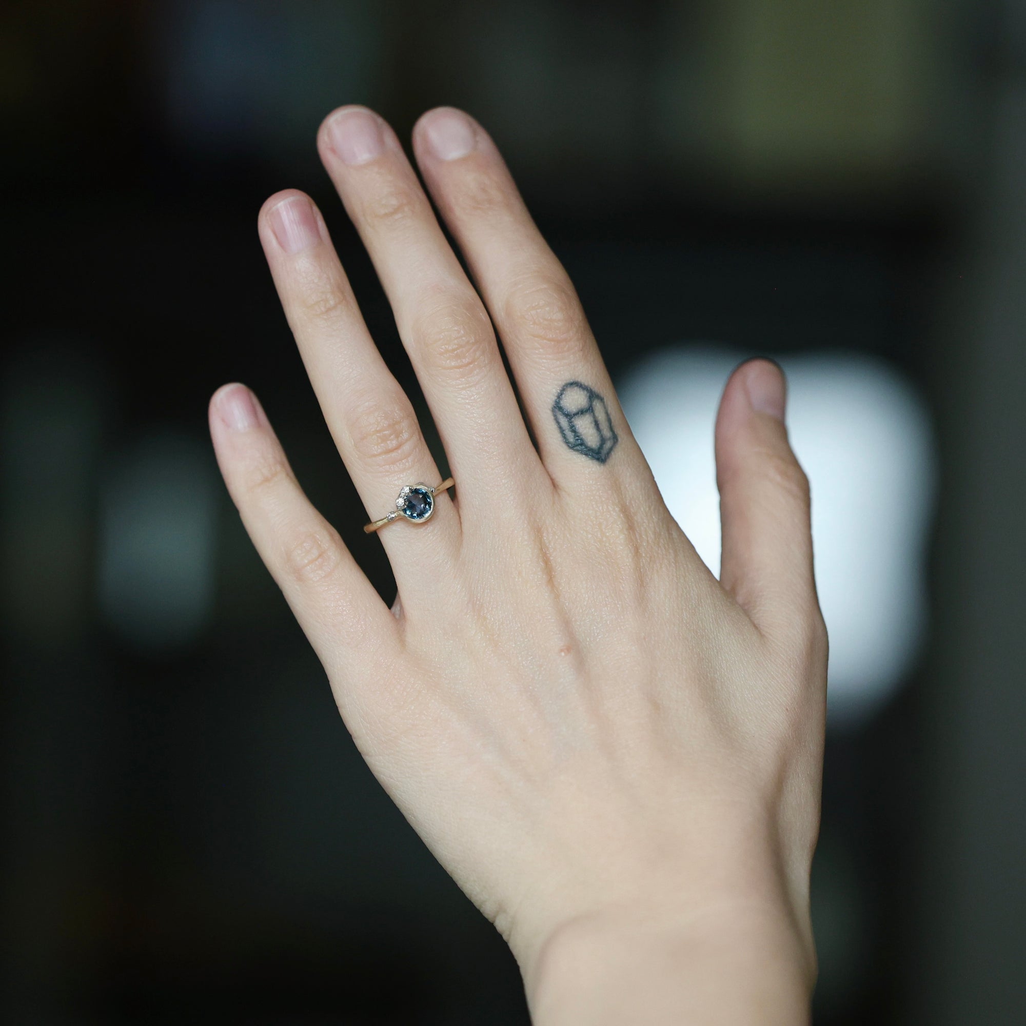A close-up of a person's hand wearing a gold ring with a dark blue gemstone, set against a dark, blurred background.
