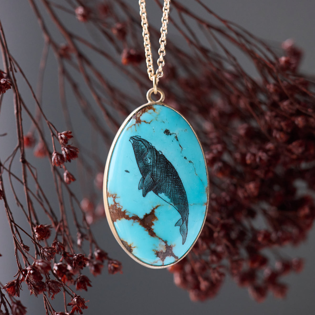 An oval pendant necklace featuring a turquoise stone with a black ink-style illustration of a whale, suspended from a gold chain against a dark gray background with blurred dried red flowers.