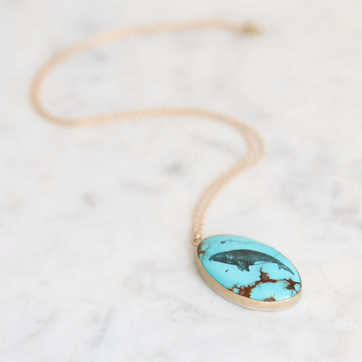 An oval turquoise pendant necklace with a gold-toned bezel and chain, resting on a white marble surface. The pendant features a black bird motif over a vibrant turquoise background with brown matrix details.