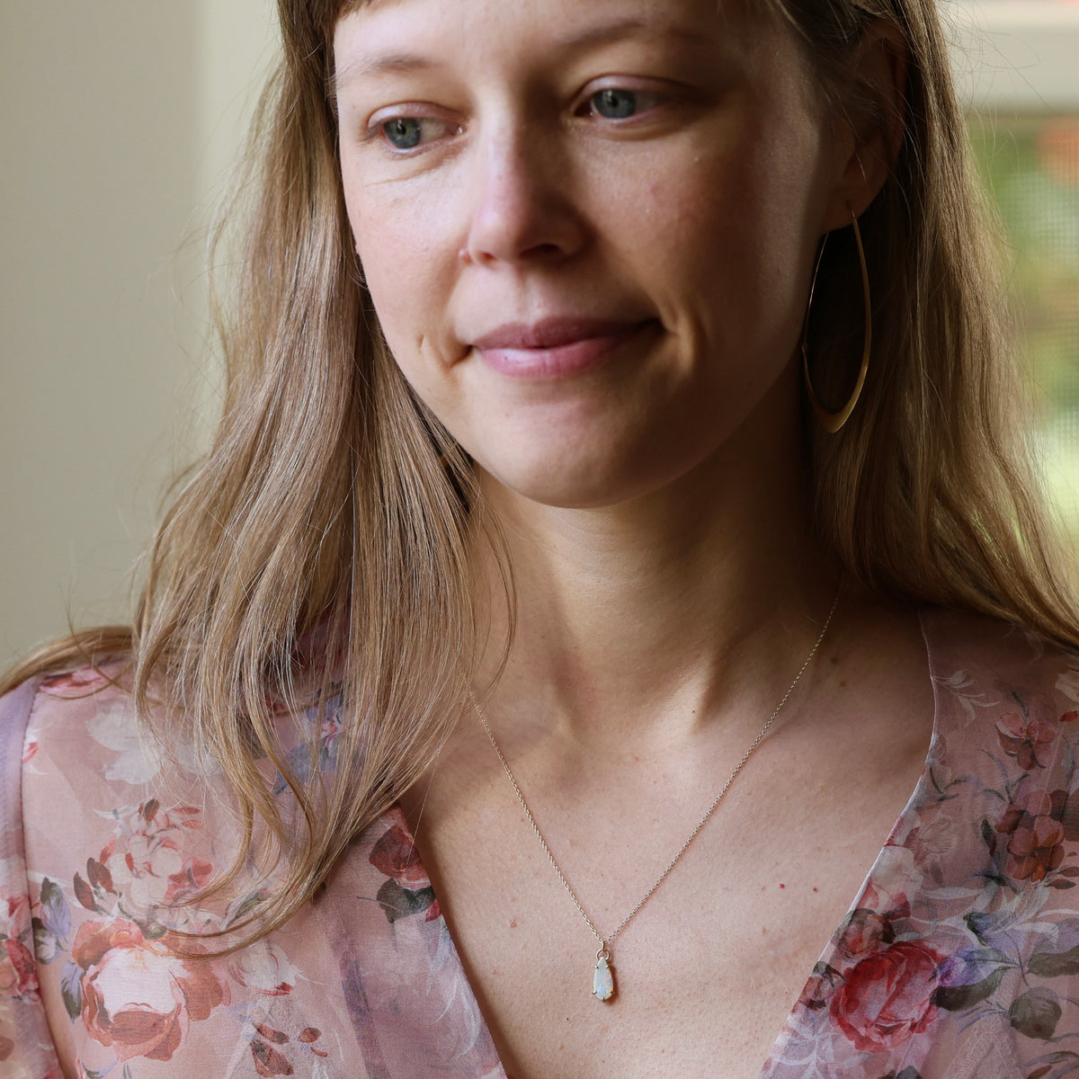 Woman wearing delicate gold chain necklace and floral blouse. She has in large gold hoops. Against a neutral indoor background. 