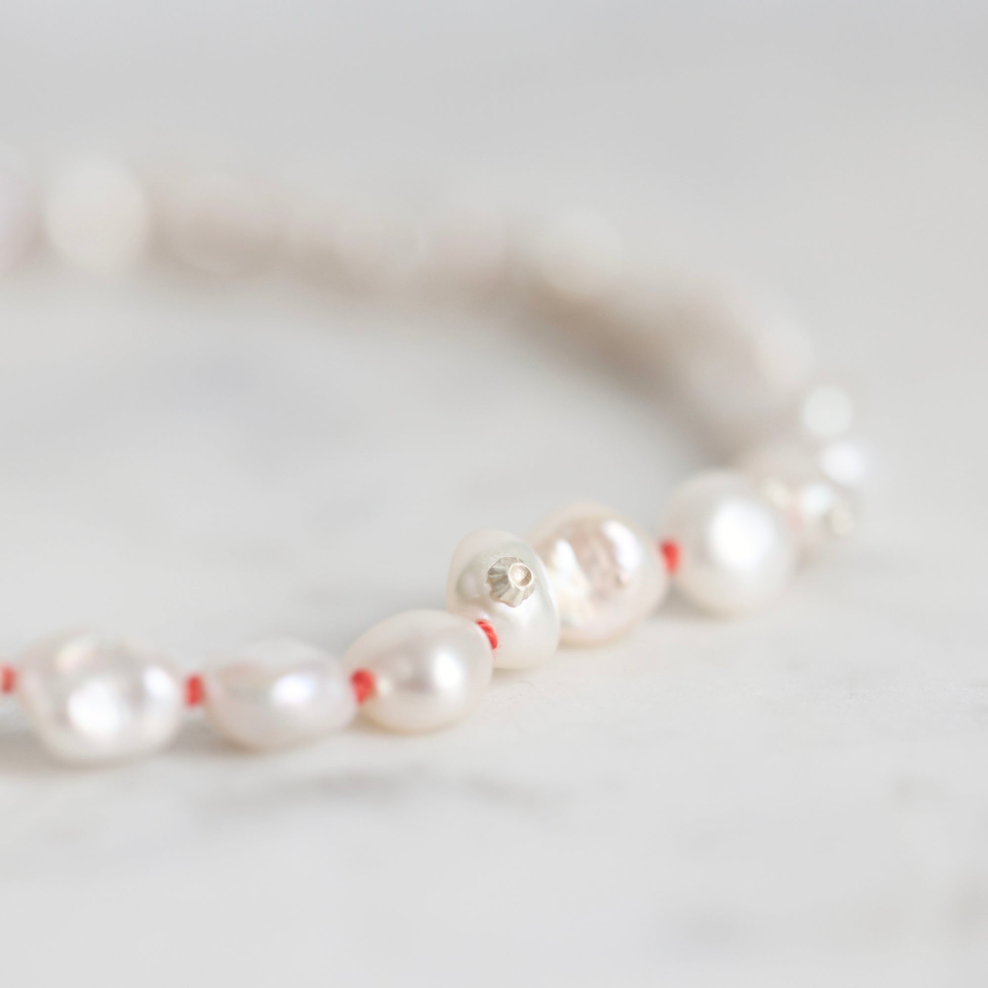 A close-up of a beaded necklace featuring lustrous white baroque pearls separated by thin red string, with a small silver-toned charm, resting on a light marble surface.