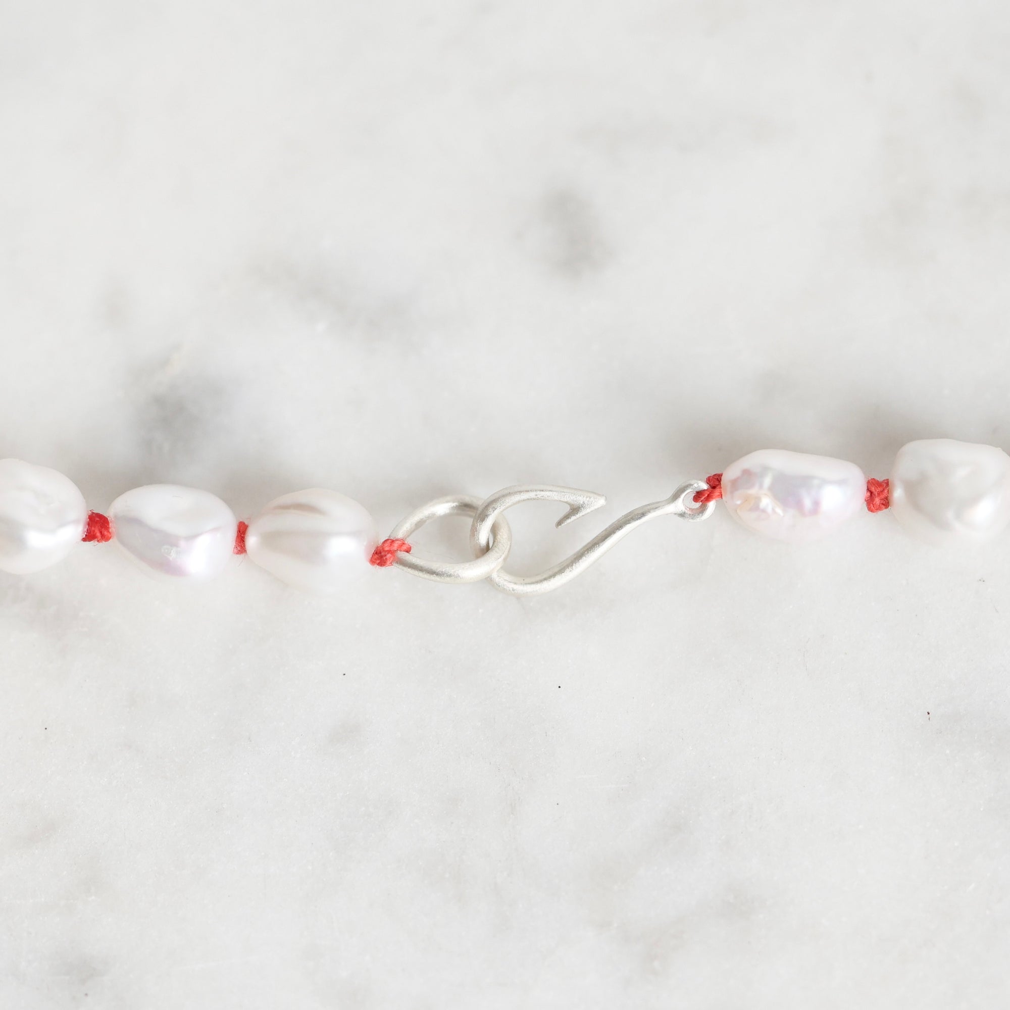 A close-up of a beaded necklace featuring irregular white baroque pearls threaded on a thin red cord, with a silver lobster clasp, set against a light gray marble background.