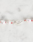 A close-up of a beaded necklace featuring irregular white baroque pearls threaded on a thin red cord, with a silver lobster clasp, set against a light gray marble background.