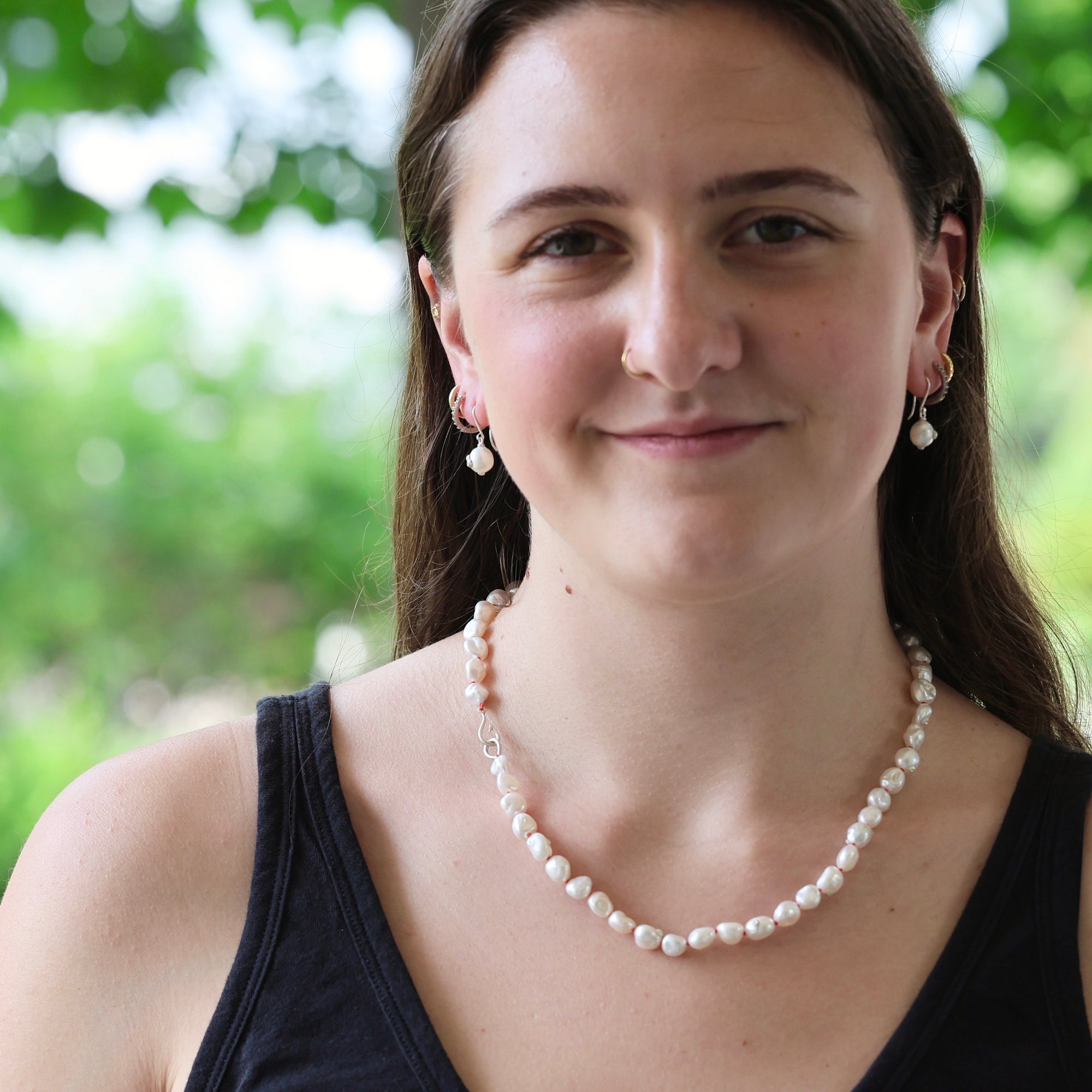 A woman with long brown hair wearing a white freshwater pearl necklace and matching pearl drop earrings, photographed outdoors with a soft green bokeh background.