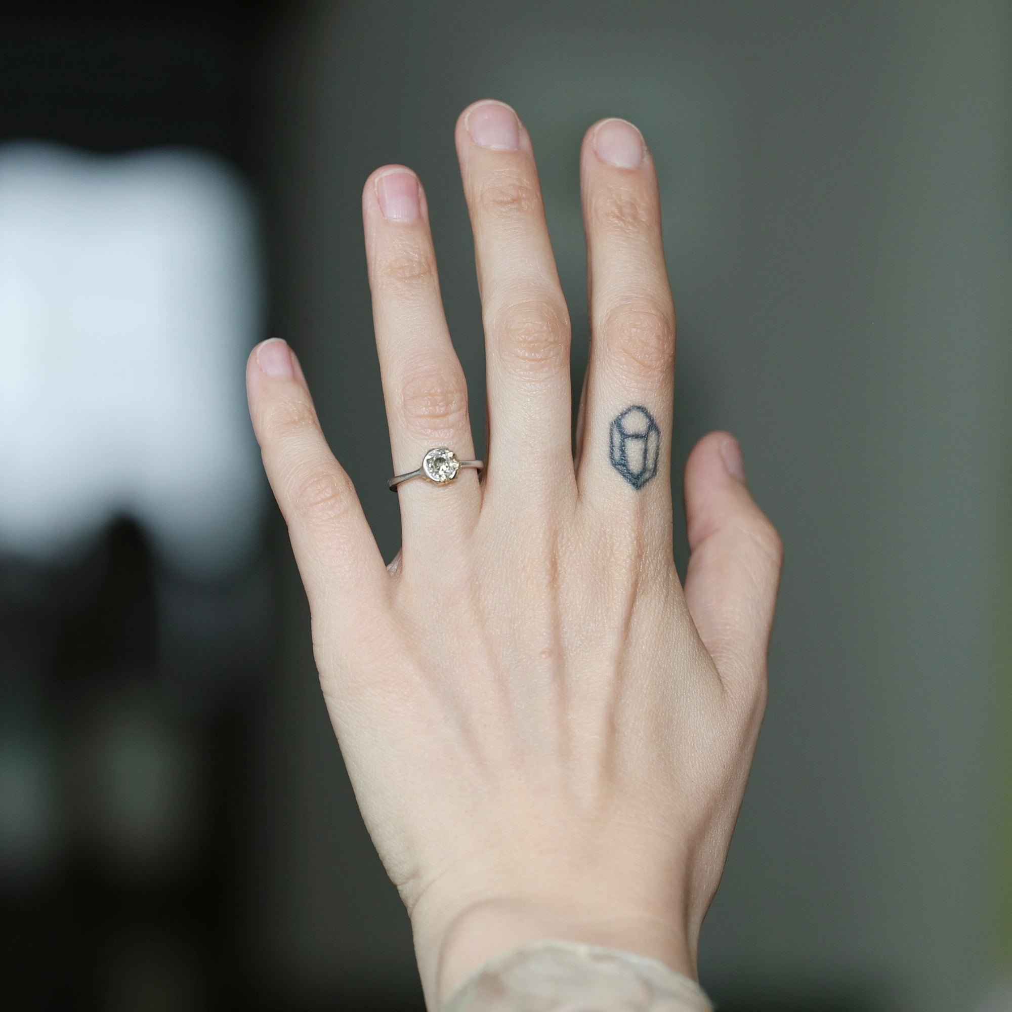 A close-up of a person's hand showing a silver ring with a round clear gemstone on the ring finger, alongside a small dark geometric tattoo on the middle finger.