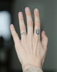 A close-up of a person's hand showing a silver ring with a round clear gemstone on the ring finger, alongside a small dark geometric tattoo on the middle finger.