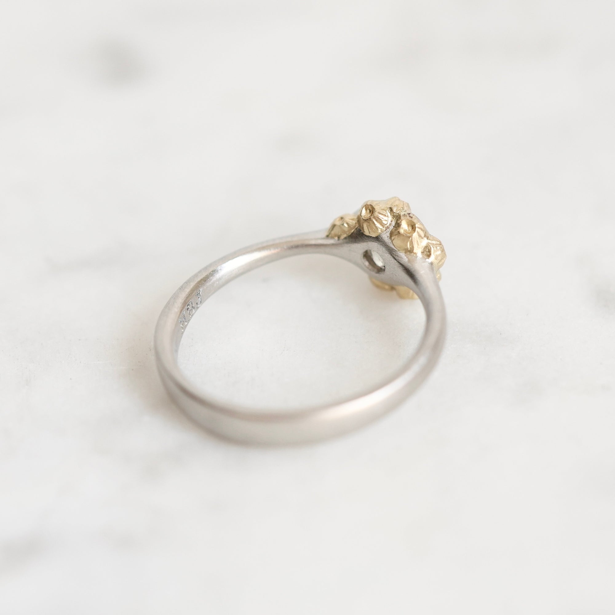 A two-tone ring featuring a polished silver band and a textured, organic-shaped gold top, resting on a light marble surface.