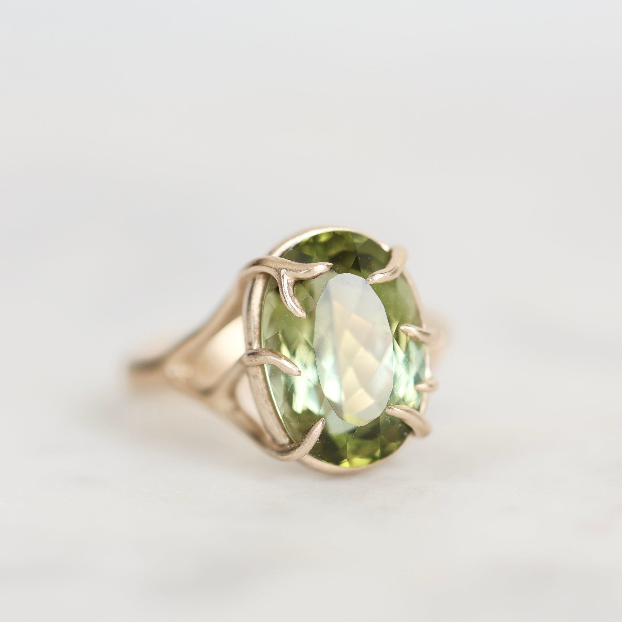 A gold ring featuring an oval-cut light green gemstone in a prong setting, photographed on a clean, light gray background.