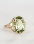 A gold ring featuring an oval-cut light green gemstone in a prong setting, photographed on a clean, light gray background.
