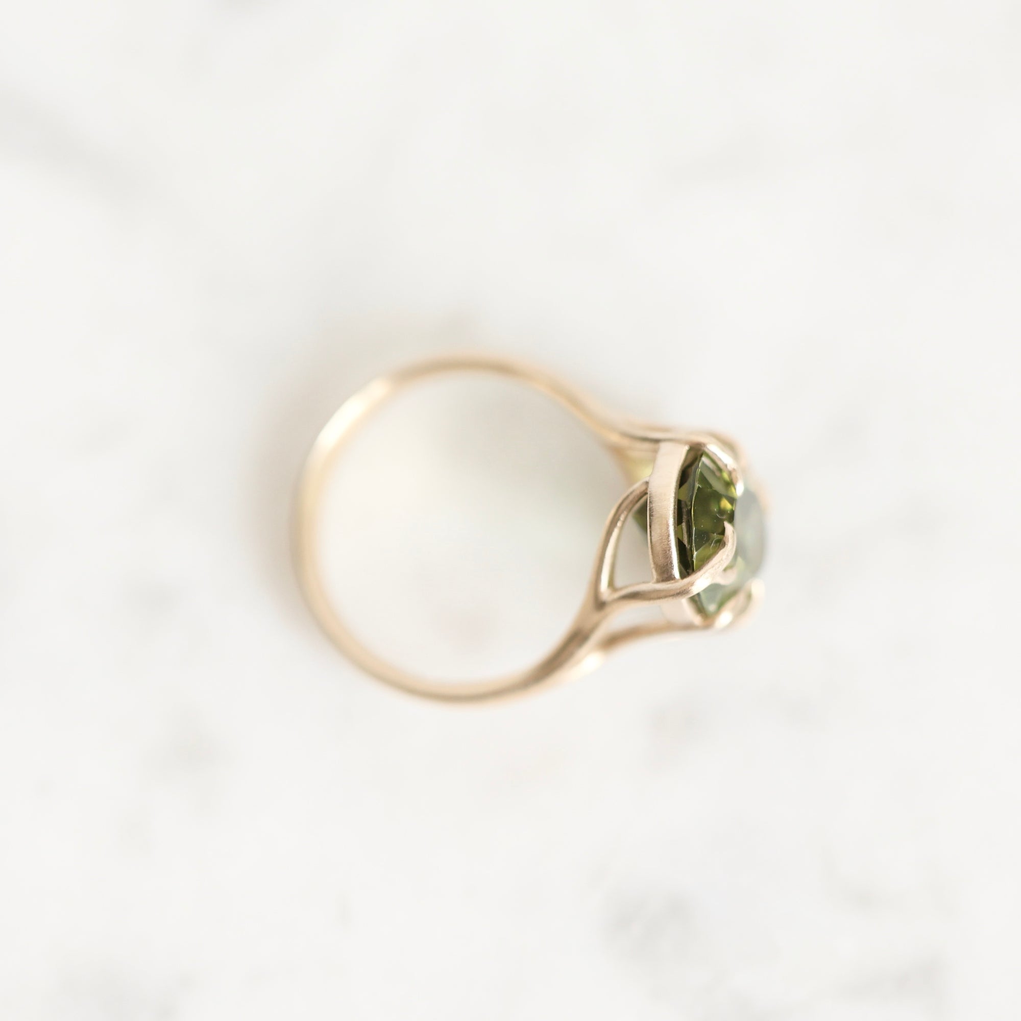 A gold ring with a marquise-cut green gemstone, photographed from above on a light gray marble surface.
