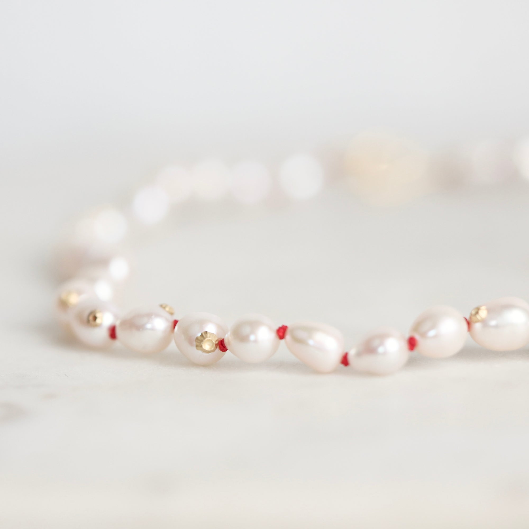 A close-up of a beaded necklace featuring irregular white pearls separated by tiny red beads and accented with small gold spacers, resting on a light marble surface.