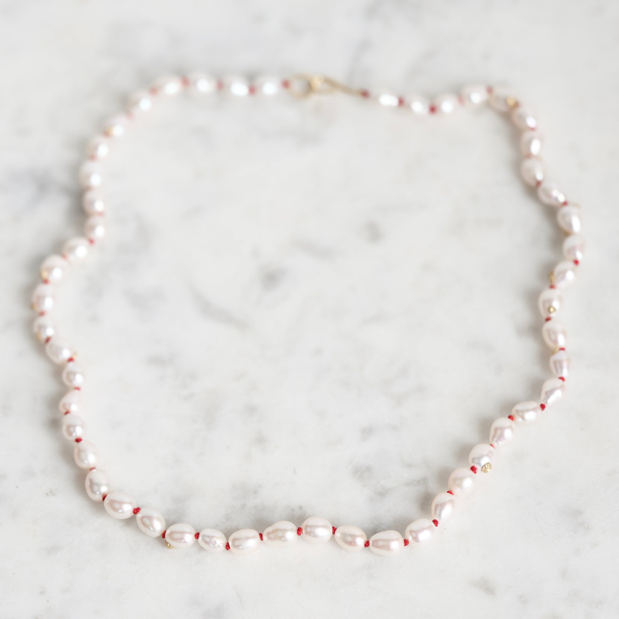 A baroque pearl necklace with small red spacer beads and a gold clasp, displayed on a light gray marble surface.