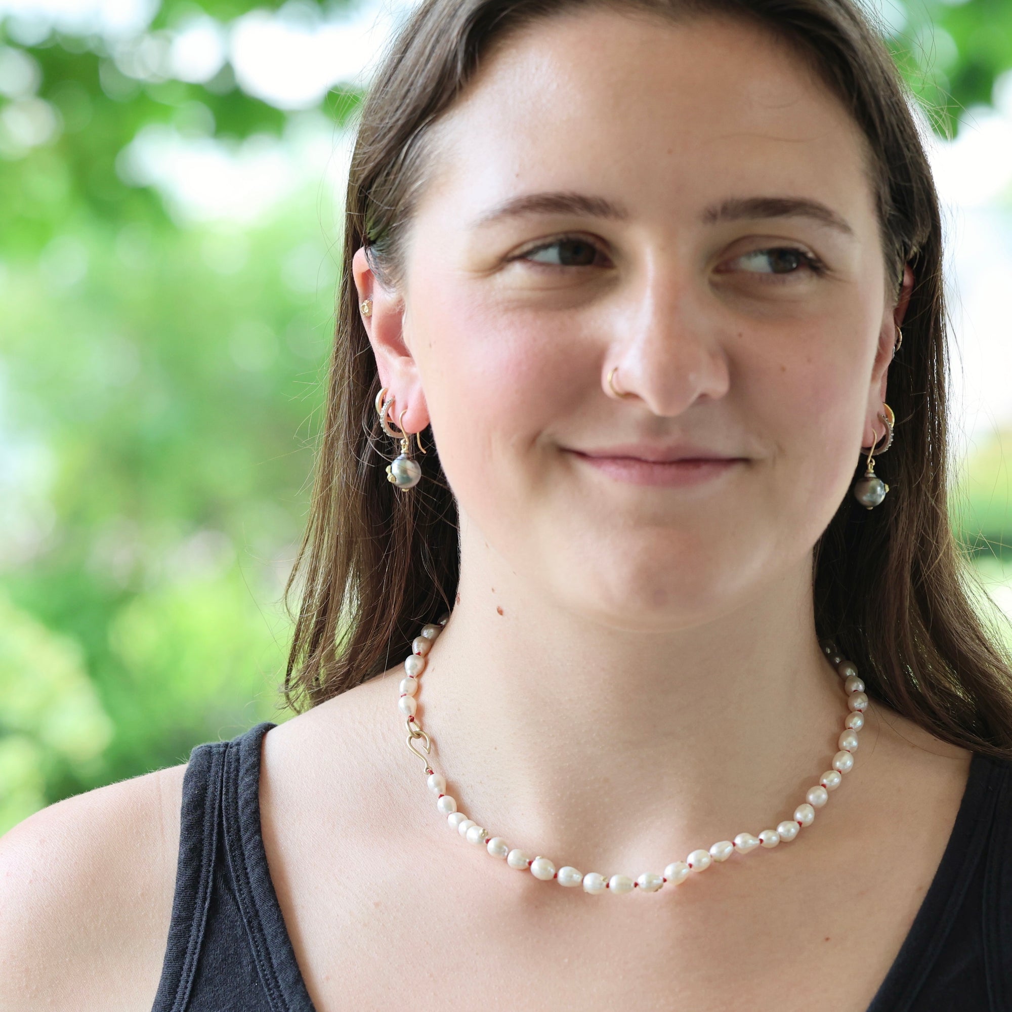 A woman wearing a delicate strand of white freshwater pearls as a necklace, styled with matching pearl drop earrings, set against a soft, blurred green outdoor background.