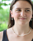 A woman wearing a delicate strand of white freshwater pearls as a necklace, styled with matching pearl drop earrings, set against a soft, blurred green outdoor background.