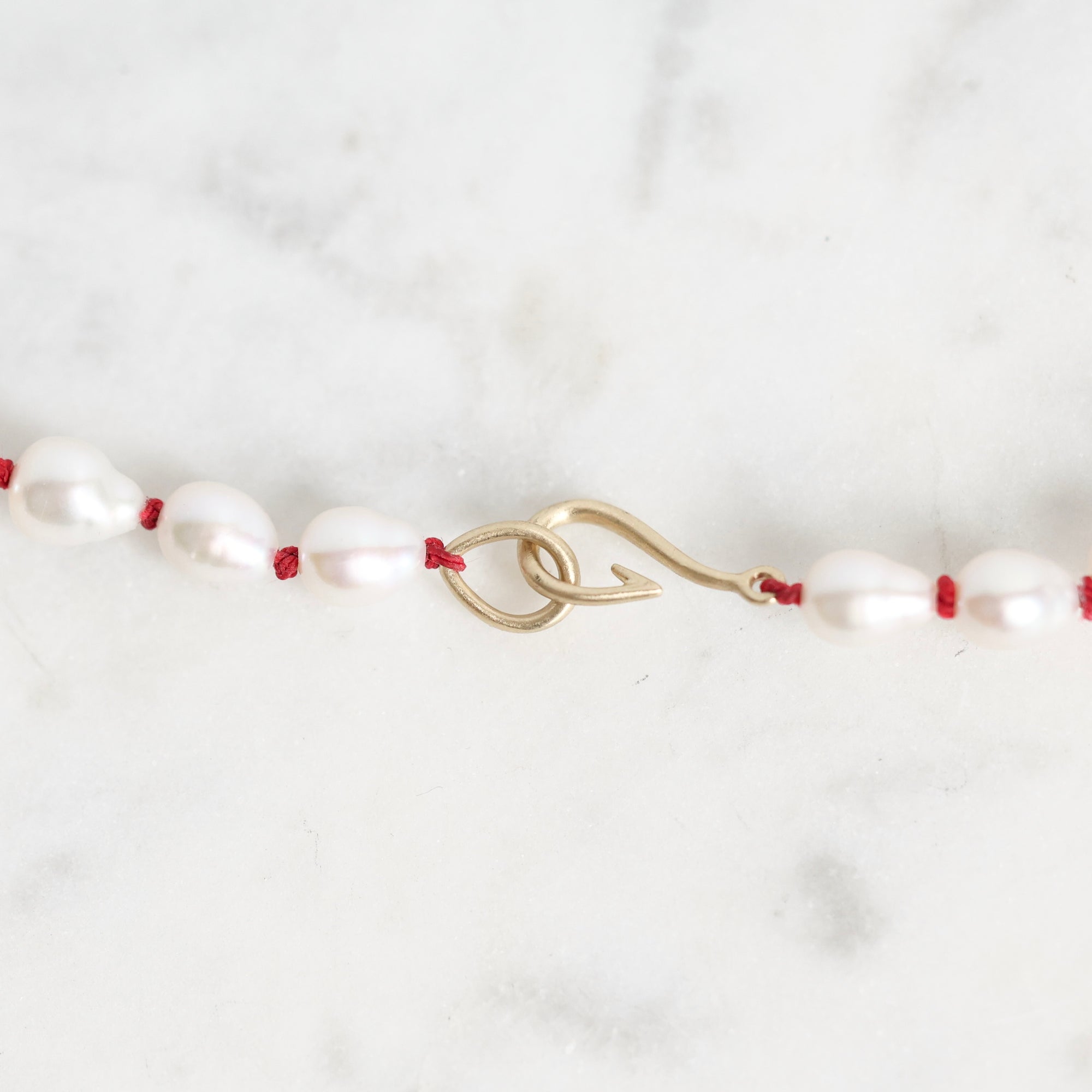 A close-up of a necklace featuring irregular white freshwater pearls strung on a thin red cord, with a gold-toned lobster clasp, set against a light gray and white marble background.
