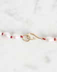 A close-up of a necklace featuring irregular white freshwater pearls strung on a thin red cord, with a gold-toned lobster clasp, set against a light gray and white marble background.