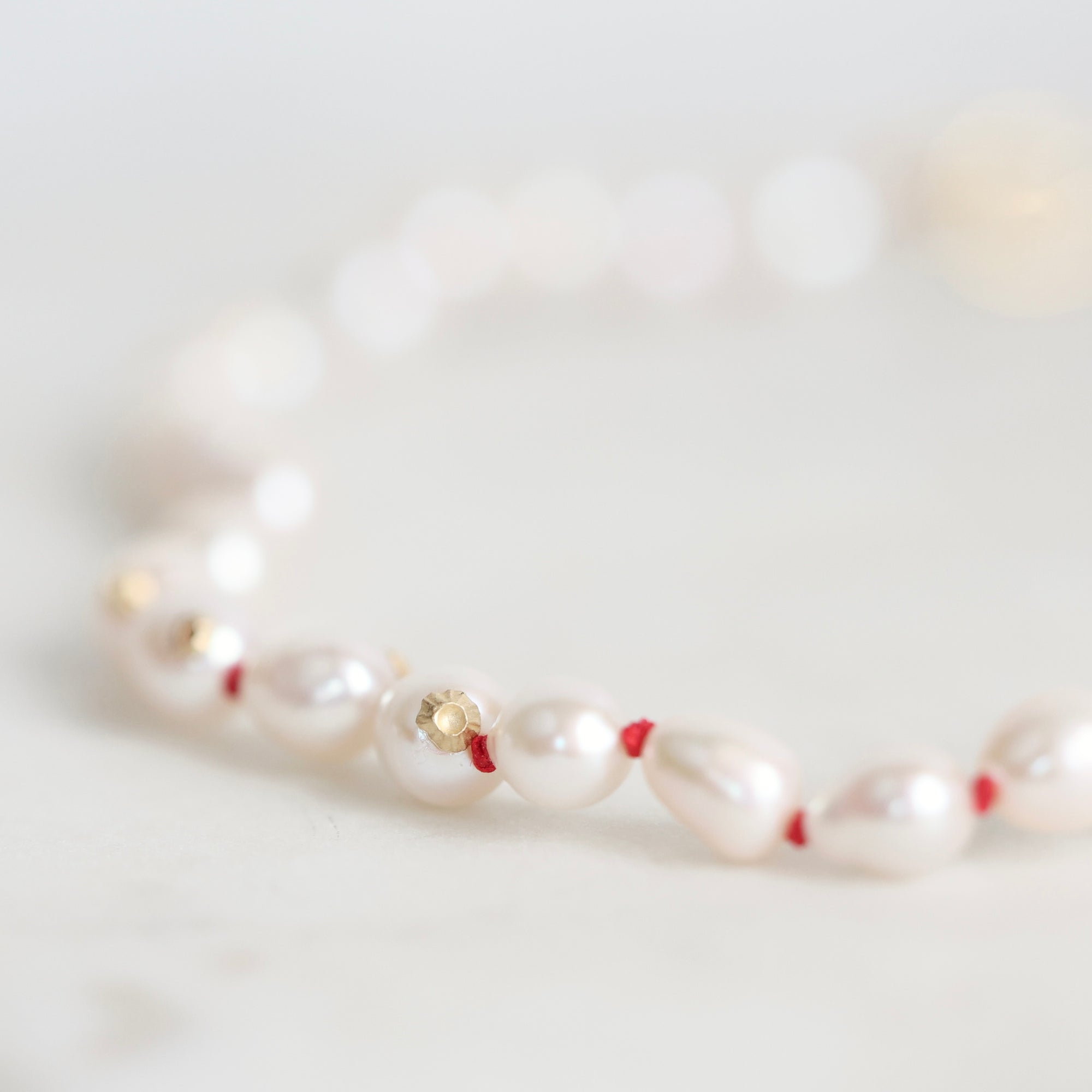 A delicate beaded bracelet featuring white baroque pearls separated by thin red string, accented with small gold spacers, photographed on a soft white surface.