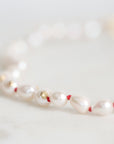 A delicate beaded bracelet featuring white baroque pearls separated by thin red string, accented with small gold spacers, photographed on a soft white surface.