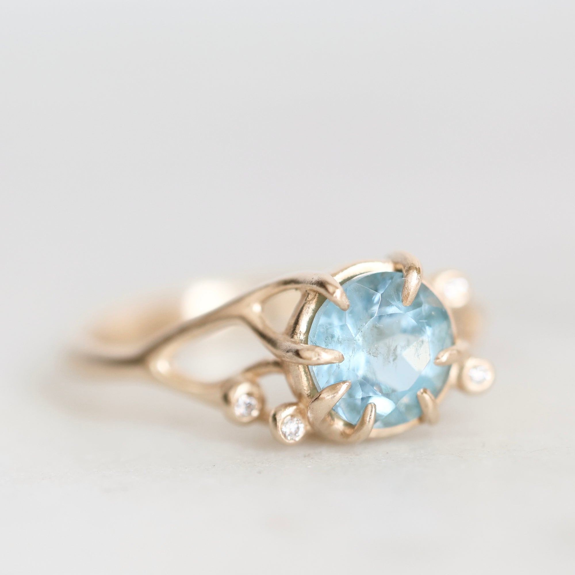 A gold ring featuring a round light blue gemstone in a prong setting, accented with small clear stones along the band, shown on a light gray background.