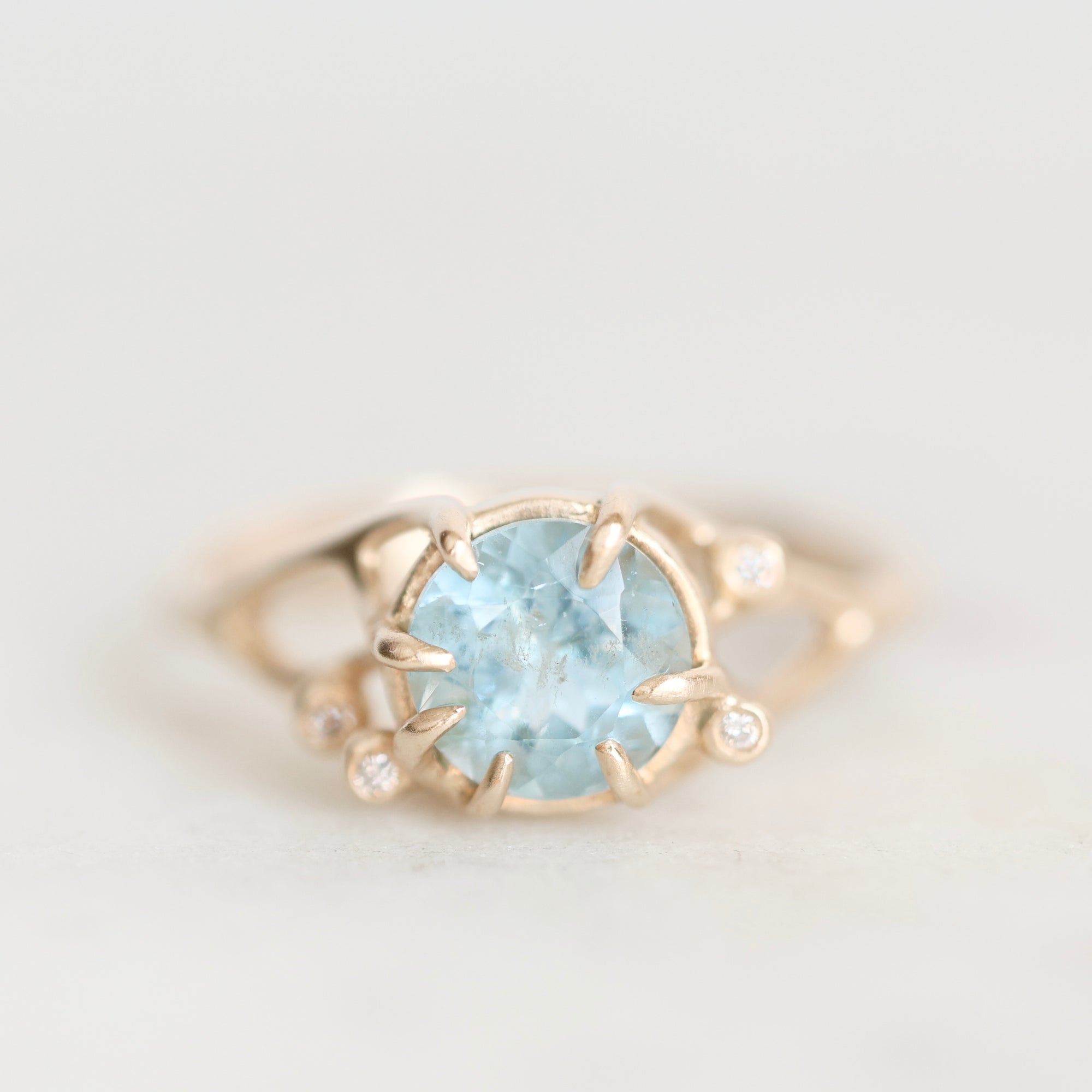 A gold ring featuring a round light blue gemstone in a prong setting, accented with small clear stones on the band, photographed against a soft light gray background.