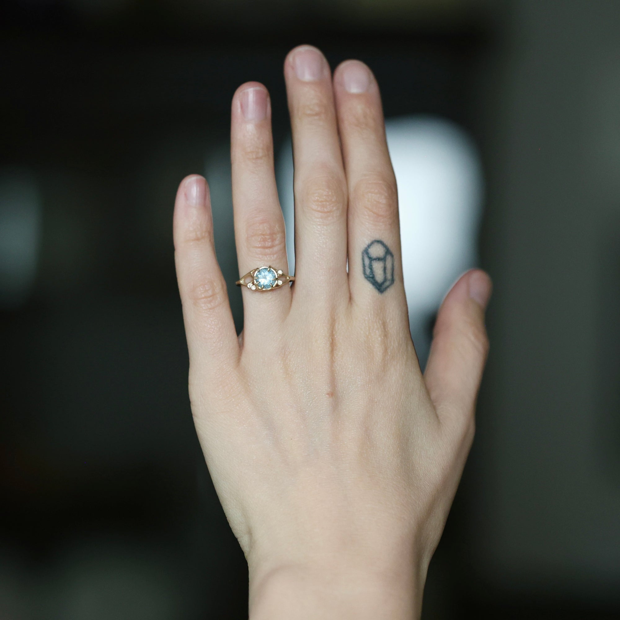 A close-up of a person's hand wearing a delicate gold ring with a round light blue gemstone, set against a dark, blurred background.