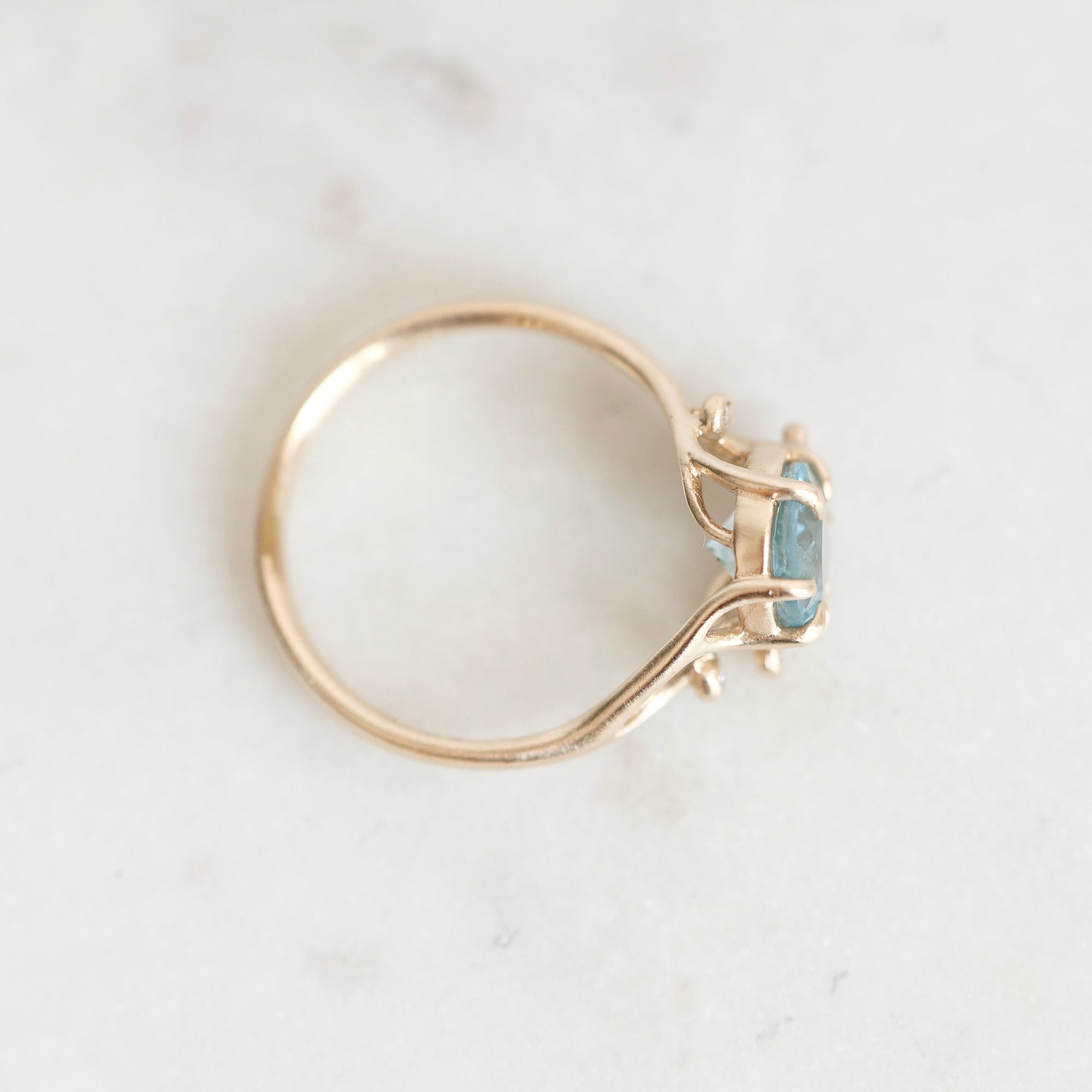 A delicate gold ring with a small light blue gemstone, photographed from above on a light marble surface.