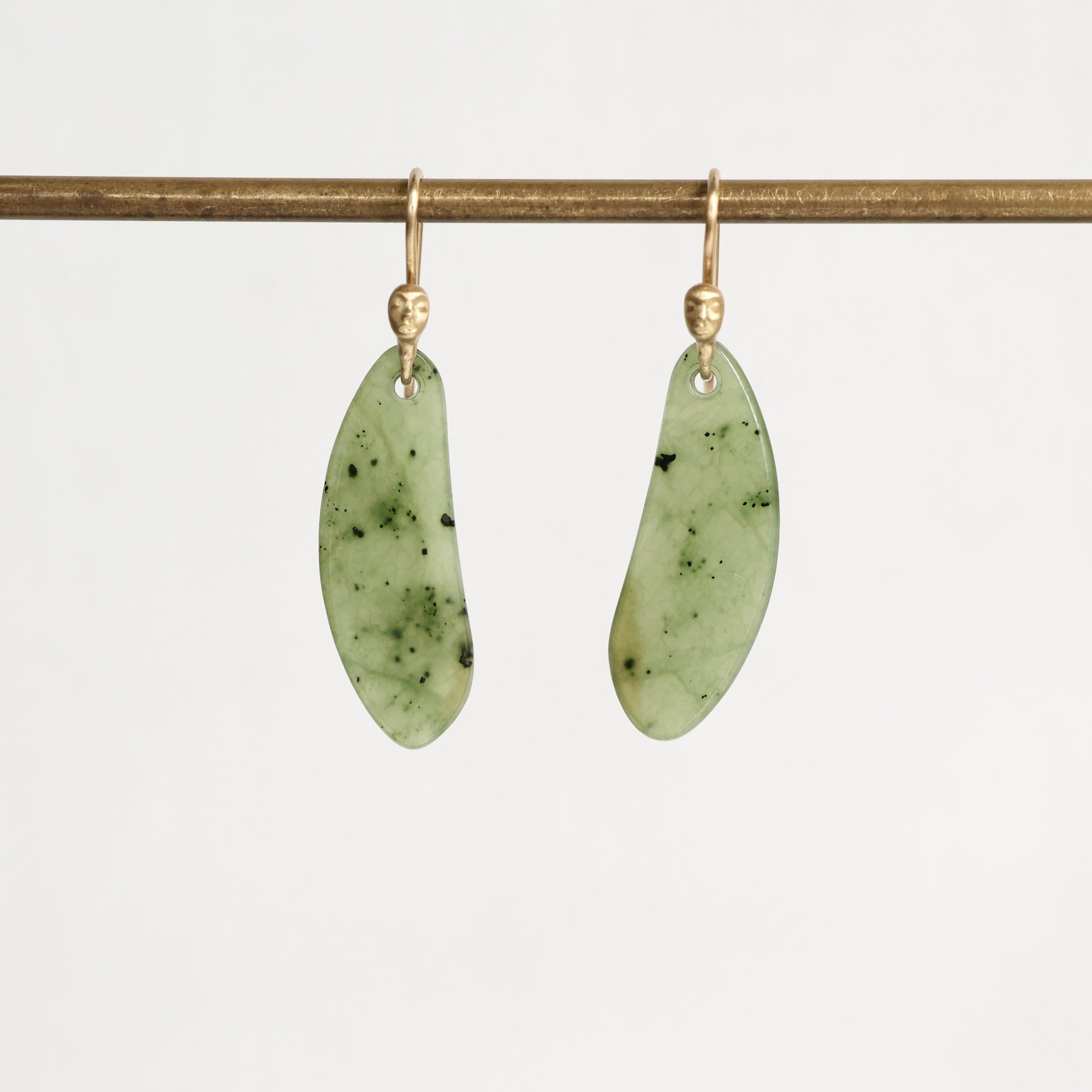 A pair of organic-shaped light green jadeite pendants with dark green inclusions, suspended from gold fishhook ear wires on a gold display bar, against a clean white background.