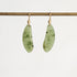 A pair of organic-shaped light green jadeite pendants with dark green inclusions, suspended from gold fishhook ear wires on a gold display bar, against a clean white background.