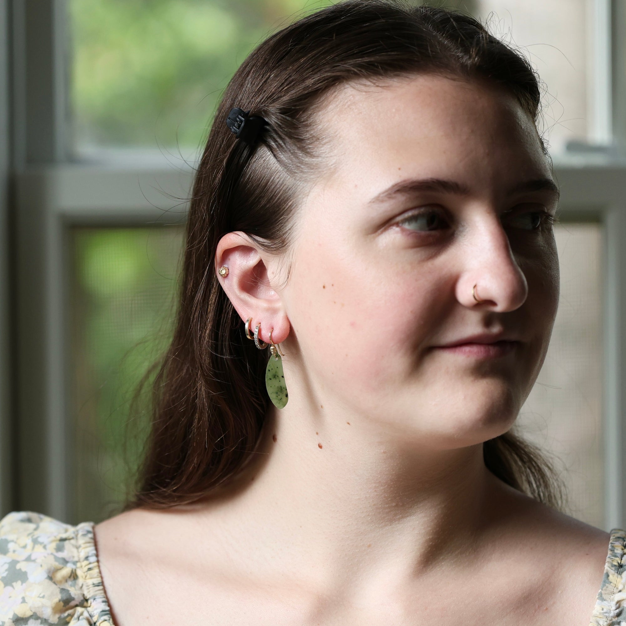 A close-up portrait of a woman with brown hair wearing a small green leaf-shaped dangle earring and multiple hoop earrings, standing in front of a softly blurred window.