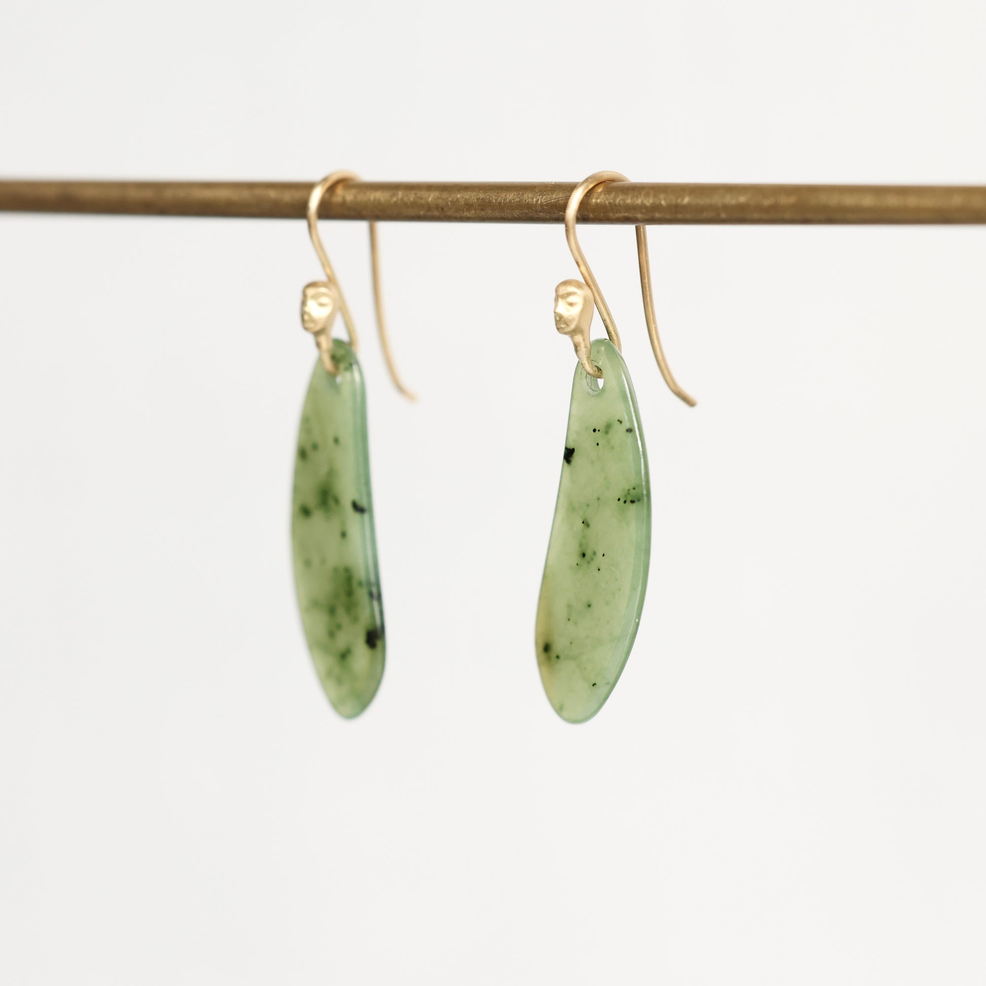A pair of gold-tone dangle earrings with elongated, polished light green stone pendants containing dark inclusions, hanging from a horizontal gold display rod against a plain white background.