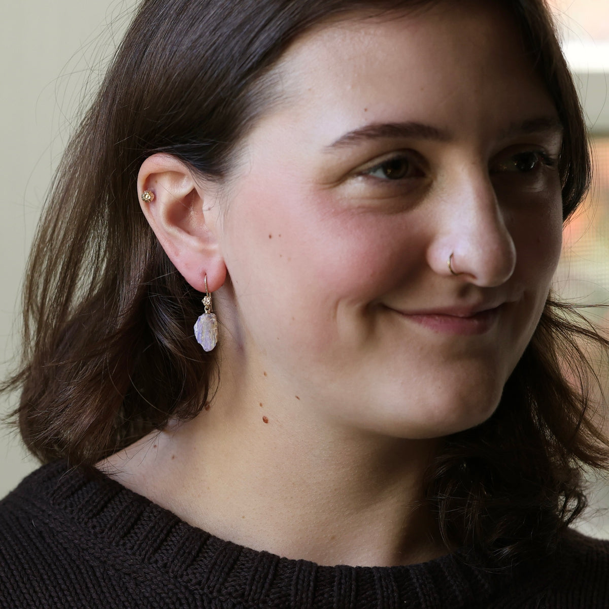 A close-up of a woman wearing a delicate gold hoop earring with a small white stone dangle, shown in profile against a softly blurred indoor background.
