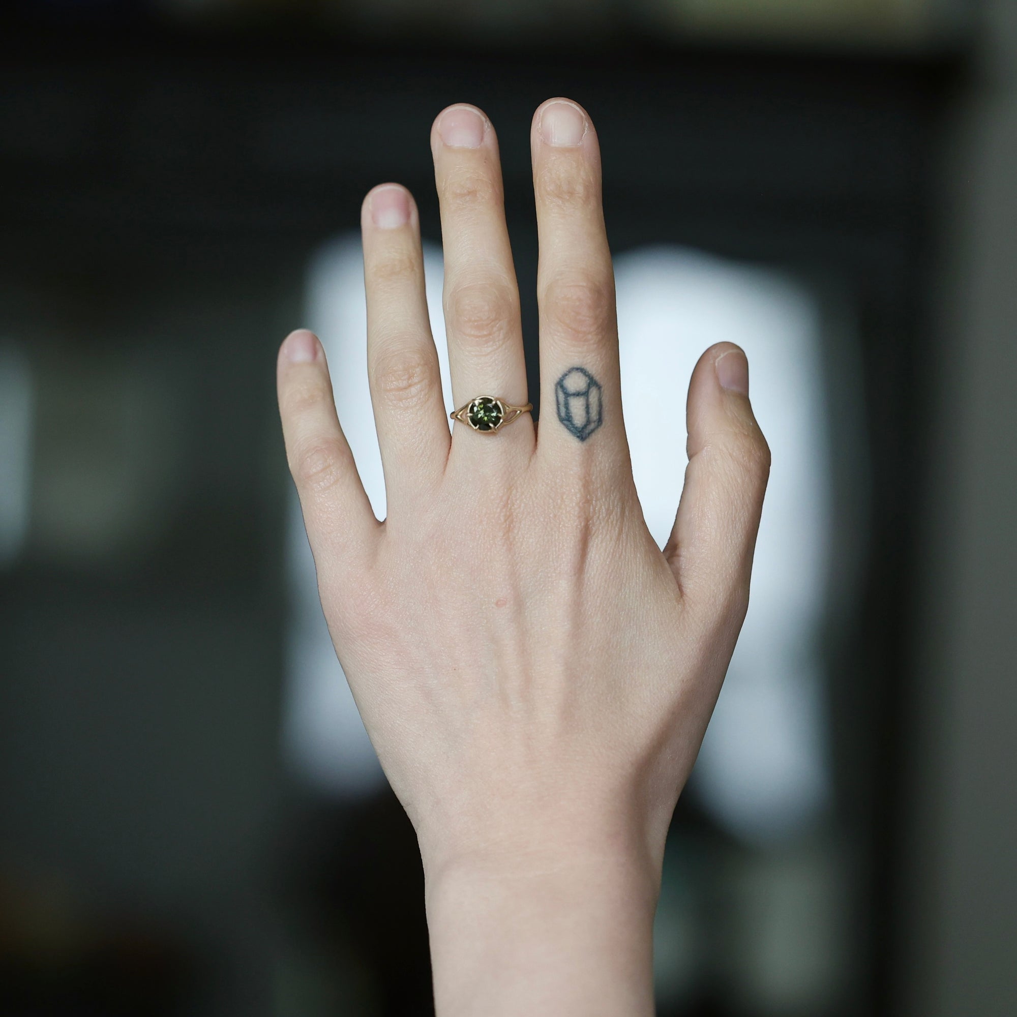 A close-up of a person's hand showing two rings: a gold ring with a small dark green gemstone on the ring finger and a silver-toned geometric ring on the middle finger, against a blurred dark background.