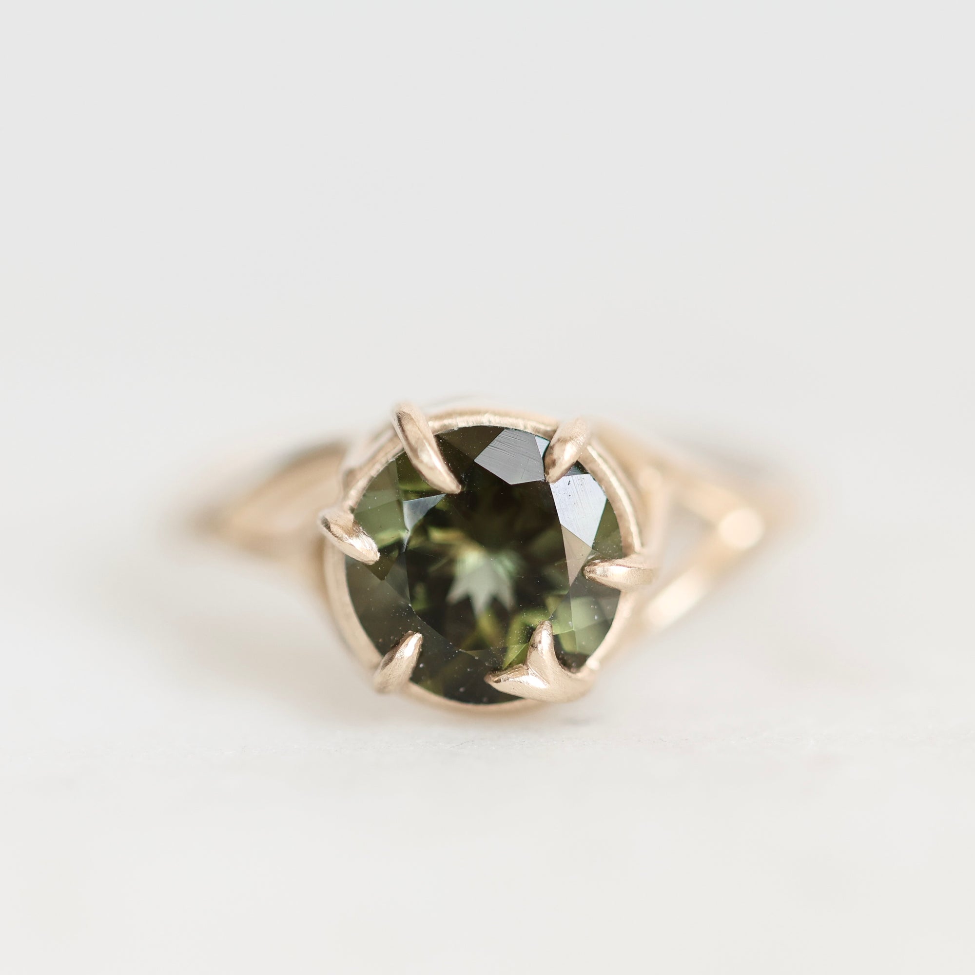 A gold ring featuring a round faceted dark green gemstone held in a prong setting, photographed on a clean white background.