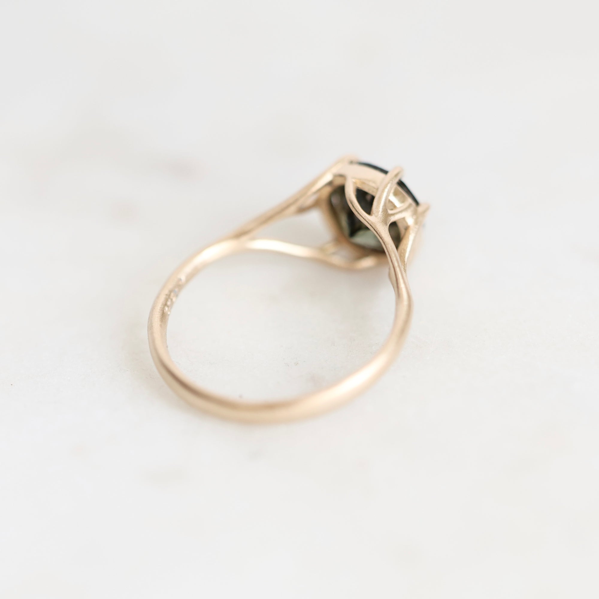 A gold ring with a split shank and a small dark green gemstone, photographed on a light gray marble surface.